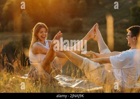 Junge Erwachsene Paare, die in der Natur Yoga praktizieren, fröhliche Frau und glücklicher Mann, die gemeinsam bei Sonnenuntergang trainieren Stockfoto
