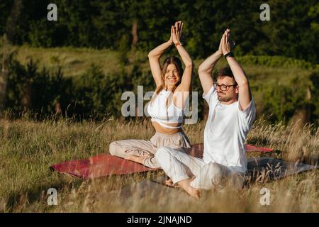 Fröhliche Frau und glücklicher Mann heben die Hände über den Köpfen, junges Paar, das im Freien in der Natur meditiert Stockfoto