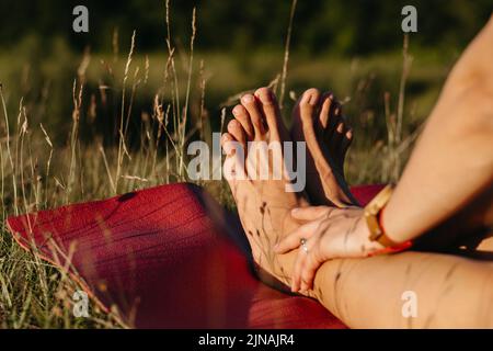 Nahaufnahme einer nicht erkennbaren Frau, die ihre Füße hält, während sie während des Yoga-Outdoors bei Sonnenuntergang auf der Matte sitzt Stockfoto