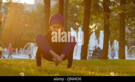 Starke islamische muslimische indische Frau in Hijab aktiv Mädchen tun Yoga im Park im Freien Training Übungen anspannen Muskeln zeigen akrobatische Elemente Stockfoto