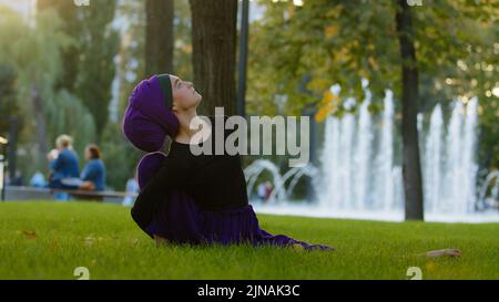 Muslimisches Mädchen in Hijab islamische Frau Yogi-Sportlerin praktiziert Yoga-Training im Park auf grünem Rasen sitzt in Garn mit einer Beinbeuge nach oben streckt Stockfoto