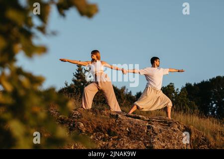 Junge Erwachsene Paare halten an den Händen, Mann und Frau üben Yoga im Freien auf dem Felsen bei Sonnenuntergang Stockfoto