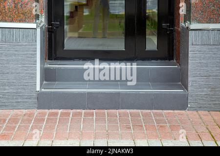 Fußgängerweg in der Nähe der Schwelle von grauen Keramikfliesen mit Stufen am Eingang zum Geschäft mit schwarzer Tür mit Glas, Fassade mit Fliesen und Grani verkleidet Stockfoto