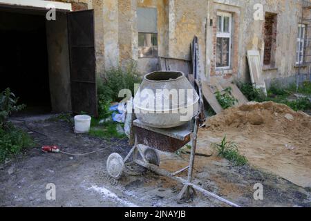 Zementmischer - Werkzeug und Gerät ist alt, alt, angeschmutzt, schmutzig, schmuddelig und verblasst. Verfallene und verfallene Gebäude, Chaos und Unordnung im Hintergrund. Stockfoto