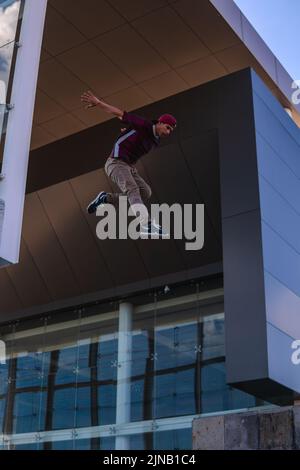 Ein Latino-Mann in städtischer Kleidung springt von der Spitze eines Gebäudes mit Architektur im Hintergrund. Hispanischer Mann macht Parkour durch Springen ich Stockfoto