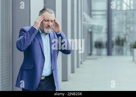 Leitender grauhaariger Geschäftsmann, krank vor dem Bürogebäude, Mann, der die Hände am Kopf hielt und schwere Migräne und Kopfschmerzen hatte Stockfoto