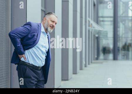 Übermüder grauhaariger älterer Mann vor dem Bürogebäude, Geschäftsmann im Geschäftsanzug, die Hand hinter dem Rücken haltend, starke Rückenschmerzen habend, lange sitzende Arbeit Stockfoto