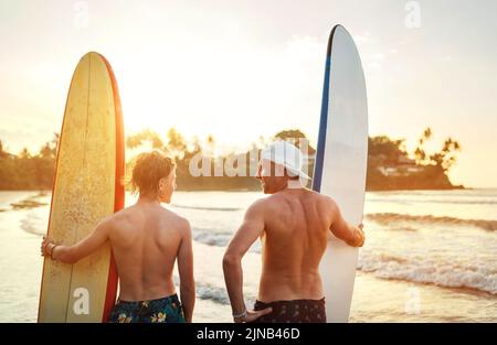 Vater mit Teenager-Sohn, der mit Surfbrettern am sandigen Meeresstrand mit Palmen im Hintergrund steht, die von der Sonne des Sonnenuntergangs erhellt werden. Sie lächeln und h Stockfoto