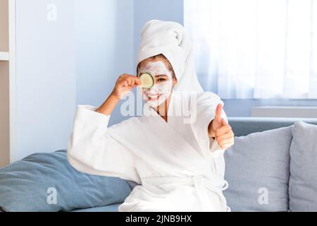 Junge Frau mit einer Gesichtsmaske aus Ton, die Gurkenscheiben hält. Schöne junge Frau mit Gesichtsmaske auf ihrem Gesicht, die Scheiben frischer Gurken hält. Haut c Stockfoto