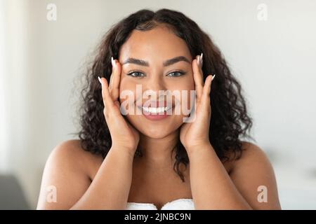 Schöne schwarze Körper positive Frau berührt ihr Gesicht und lächelt an der Kamera, machen Schönheitsbehandlungen zu Hause Stockfoto