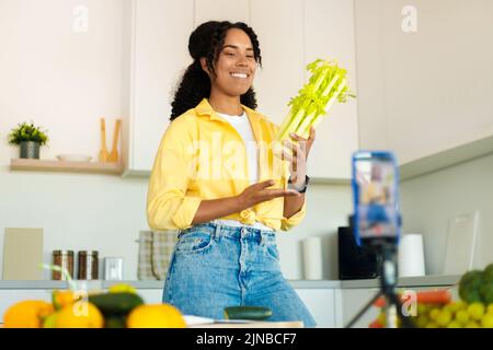 Die junge schwarze Dame filmte Inhalte in der Küche für ihren Food-Blog, benutzte ein Smartphone auf einem Stativ und zeigte Sellerie vor der Kamera Stockfoto