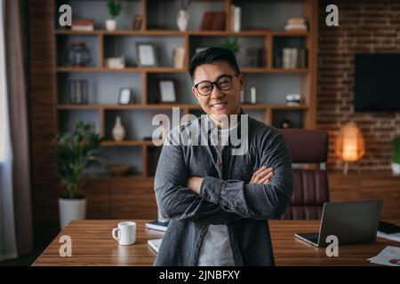 Lächelnder, selbstbewusster, erfolgreicher erwachsener chinese in einer Brille mit gekreuzten Armen auf der Brust und Blick auf die Kamera Stockfoto