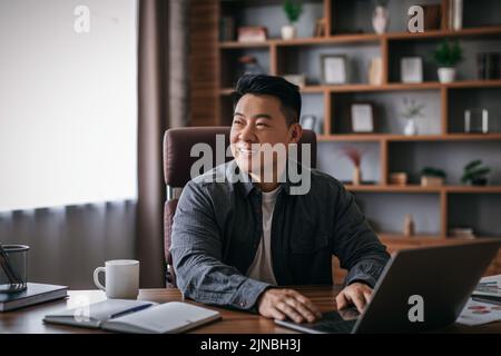 Glücklicher reifer chinese am Tisch, der mit einem Laptop im Home Office-Innenraum arbeitet und auf leeren Raum schaut Stockfoto
