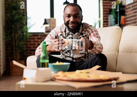 afroamerikanischer Mann, der die fernbedienung des fernsehers benutzte, um den Sender zu wechseln und ein Stück Pizza von der Lieferung mit Schnellimbiss zu essen. Spaß mit Essen zum Mitnehmen und Bier, während er sich einen Film zusieht. Stockfoto