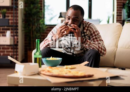 Fröhlicher Erwachsener, der zu Hause vor dem Fernseher ein Stück Pizza isst und mit einer Flasche Bier zum Mitnehmen zubereitet. Filme im Fernsehen ansehen und zum Abendessen Fastfood zum Mitnehmen genießen. Stockfoto