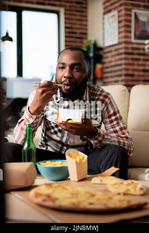 Entspannter Mann, der vor dem Fernsehfilm Nudeln vom Take-away-Lieferservice genießt und Essen mit Essstäbchen isst. Film auf dem fernsehprogramm ansehen, Spaß mit Essen zum Mitnehmen haben. Stockfoto