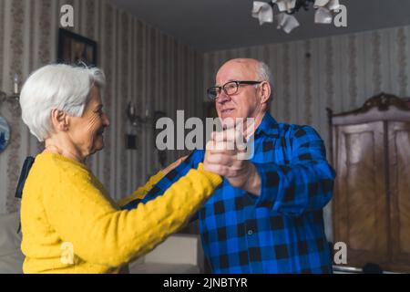 Glückliches, romantisches altes Ehepaar, das im Wohnzimmer lächelt und tanzt, die Hände hält und Spaß zu Hause hat. Hochwertige Fotos Stockfoto