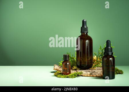 Ein Set brauner Glasflaschen, Moos und Baumrinde auf grünem Hintergrund. Öl mit Serum zur Haut- und Haarpflege. Glasflasche mit ätherischem Körperöl Stockfoto