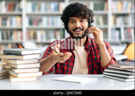 Ein positiver arabischer oder indischer Student mit lockigen Haaren, mit einem Headset, sitzt mit Büchern an einem Tisch in der Bibliothek, schaut sich ein Online-Webinar an, hört sich einen Vortrag an, schaut lächelnd auf die Kamera Stockfoto