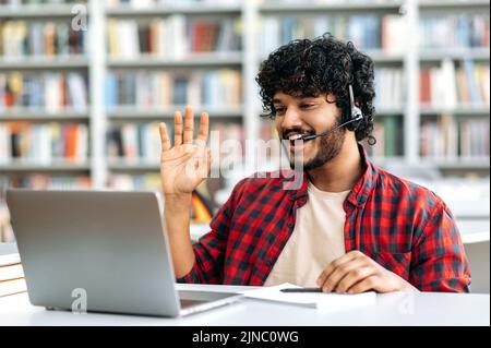 Fröhlicher, moderner Inder oder Araber, Student oder Freiberufler mit Headset, sitzt an einem Schreibtisch in einer Bibliothek, nutzt Laptop für Videoanruf, winkt, hört Online-Webinar, Vortrag, lächelt Stockfoto