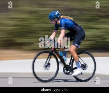 Eine Radfahrerin an der Spitze, die auf einer Runde eines Criterium ihr Fahrrad fährt, ein schnelles Radrennen um wiederholte Kurzstrecken auf den Straßen der Stadt. Bewegungsunschärfe. Stockfoto