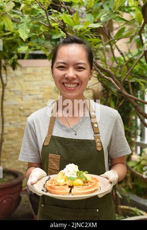 Die vietnamesische Kellnerin servierte belgische Waffeln mit Obst und Eis im Café im Freien und schaute nach oben Stockfoto