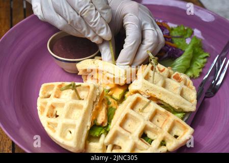 Hände in Gummihandschuhen, die belgische Waffeln mit gebratenem Huhn auf violettem Teller servieren Stockfoto