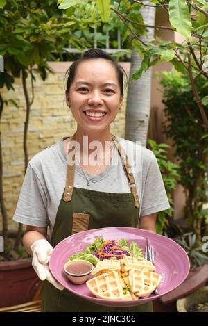 Vietnamesische Kellnerin serviert belgische Waffeln mit Gemüse und Huhn im Café im Freien Stockfoto