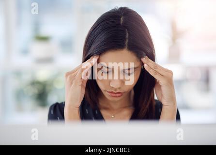 Es gibt zu viel zu tun und so wenig Zeit. Eine attraktive junge Geschäftsfrau, die an Kopfschmerzen in ihrem Büro bei der Arbeit leidet. Stockfoto