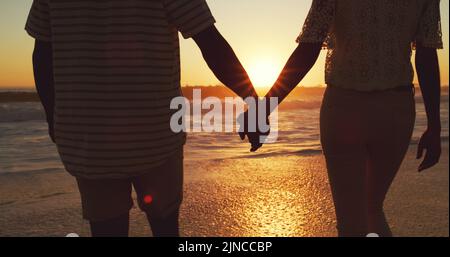 Unsere Liebe ist tiefer als der Ozean. Rückansicht eines nicht erkennbaren jungen Paares, das bei Sonnenuntergang die Hände am Strand hält. Stockfoto