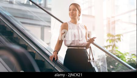 Shes zufrieden mit, wie weit shes kommen. Eine attraktive junge Geschäftsfrau, die nachdenklich aussieht, während sie an einem modernen Arbeitsplatz eine Rolltreppe hinuntergeht. Stockfoto