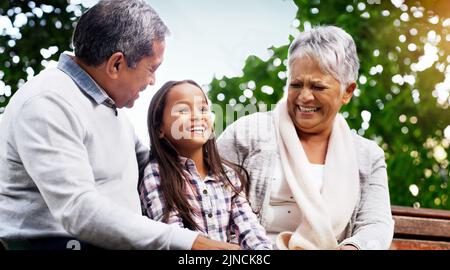 Sie erhellt immer unseren Tag. Ein fröhliches kleines Mädchen, das mit ihren Großeltern im Park auf einer Bank sitzt. Stockfoto