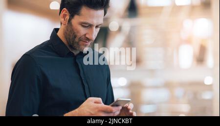 Ein gutaussehender Geschäftsmann mittleren Alters, der ein Smartphone benutzt, während er an einem modernen Arbeitsplatz steht. Stockfoto