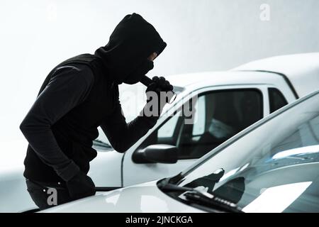 Der Raubtier auf dem Parkplatz. Ein maskierter Verbrecher, der mit einer Taschenlampe in ein leeres Auto auf einem Parkplatz schaut. Stockfoto