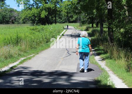 Alte Frau mit langen Haaren, die im Sommer auf dem North Branch Trail in Miami Woods in Morton Grove, Illinois, spazierengeht Stockfoto