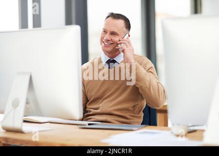 Ein gutaussehender Geschäftsmann mittleren Alters, der in einem modernen Büro an einem Computer arbeitet und einen Telefonanruf abnimmt. Stockfoto