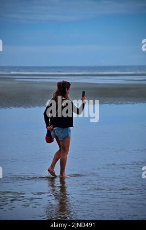 Reife Frau, die am schönen Strand spazieren geht und das Handy benutzt Stockfoto