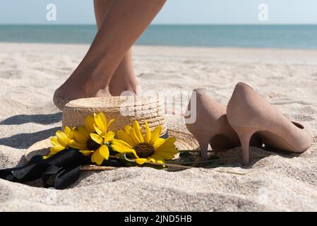 Strohhut mit Sonnenblumen, die am Meer liegen, beige Wildlederstletten, die in der Nähe stehen, und Beine einer barfuß wandenden Frau im Meersand, selektiver Fokus. Stockfoto