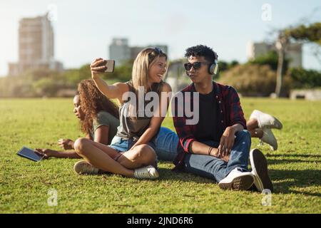 Zwei fröhliche junge Freunde, die zusammen ein Selbstporträt machen, während sie draußen in einem Park Musik auf Kopfhörern hören. Stockfoto