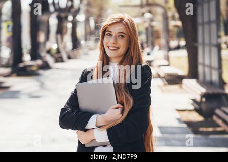 Lächelnde Ingwer-Frau schaut weg, trägt Laptop wartet auf jemanden im Park. Kaukasische Frau, die am Laptop arbeitet, freiberuflich Stockfoto