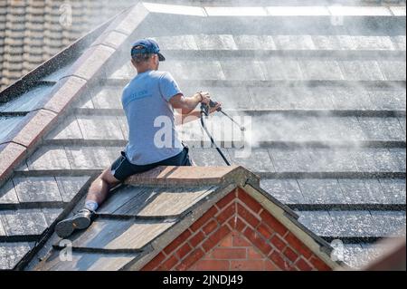 Mann sitzt auf einem Ziegeldach eines Hauses, benutzt einen Hochdruckreiniger an einem Schlauch, feuert einen Wasserstrahl ab, um das Dach und die Fliesen zu reinigen. Stockfoto