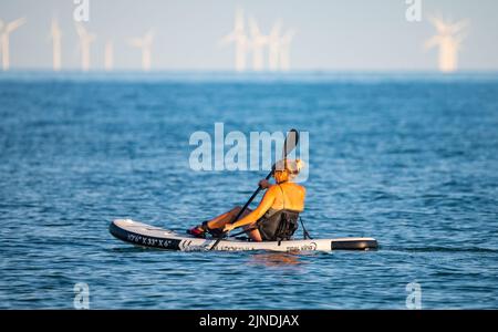 Frau sitzt auf einem Paddleboard und paddelt an einem heißen Tag im Sommer in England, Großbritannien, mit Windturbinen von einer Offshore-Windfarm im Hintergrund. Stockfoto