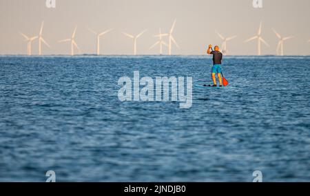 Ein Mann steht auf dem Paddleboard und paddelt an einem heißen Tag im Sommer in England, Großbritannien, mit Windturbinen von einer Offshore-Windfarm im Hintergrund. Stockfoto