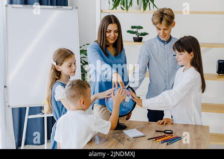 Berühren Sie den Erdball. Eine Gruppe von Kindern unterschiedlichen Alters steht in einem geräumigen Klassenzimmer in der Schule, wobei der Lehrer den Globus berührt. Stockfoto