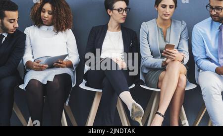 Geschäftsleute, die Schlange stehen und Informationen mit Technologie teilen. Profis sitzen in einer Reihe für ein Interview vor grauem Hintergrund. A Stockfoto