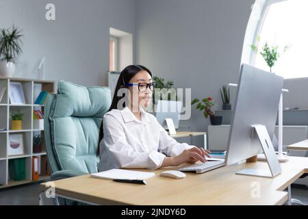Online-Fernunterricht. Junge schöne asiatische Lehrerin mit Kopfhörern und Brille sitzt an einem Tisch an einem Computer und unterrichtet online eine Klassenstunde. Stockfoto