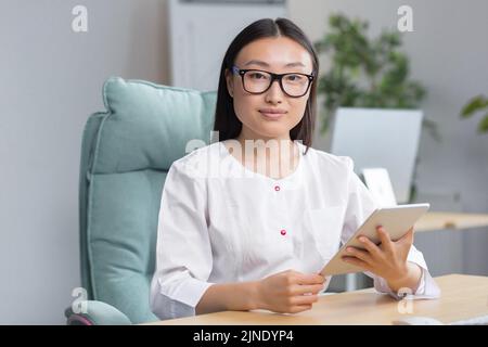 Online-Aufnahme ins Krankenhaus. Eine junge, schöne asiatische Ärztin zeichnet die Krankengeschichte eines Patienten online auf einer Tablette auf. Tippen Sie auf ein Tablet. Sitzen am Tisch im Büro in der Winding. Stockfoto