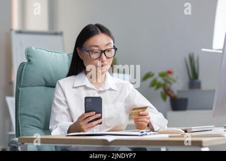 Online-Shopping. Eine junge, schöne asiatische Frau hält ein Mobiltelefon und eine Kreditkarte in der Hand, macht eine Bestellung am Telefon, zahlt per Karte. Sitzen an einem Tisch in einem modernen Büro. Stockfoto