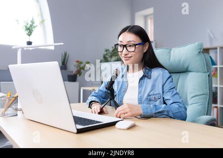 Podcast-Aufzeichnung, Interview. Junge schöne asiatische Frau, die mit einem Mikrofon und einem Laptop an einem Tisch sitzt. Spricht, erzählt. Macht eine Audioaufnahme eines Podcasts, gibt ein Interview per Videoanruf. Stockfoto