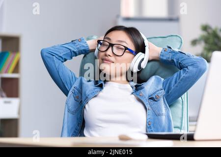 Junge, schöne asiatische Frau, die Musik über Kopfhörer hört, die Augen schließt und ihre Hände hinter ihren Kopf wirft, ruht auf einem Nahaufnahme-Foto Stockfoto